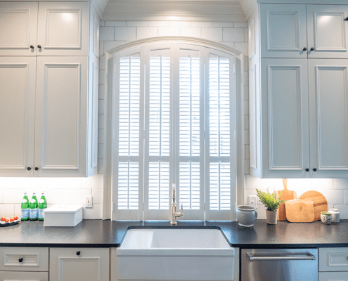 White interior shutter sets built by Dwell Shutter & Blinds displayed in a beautiful kitchen with white cabinets and backsplash tile.
